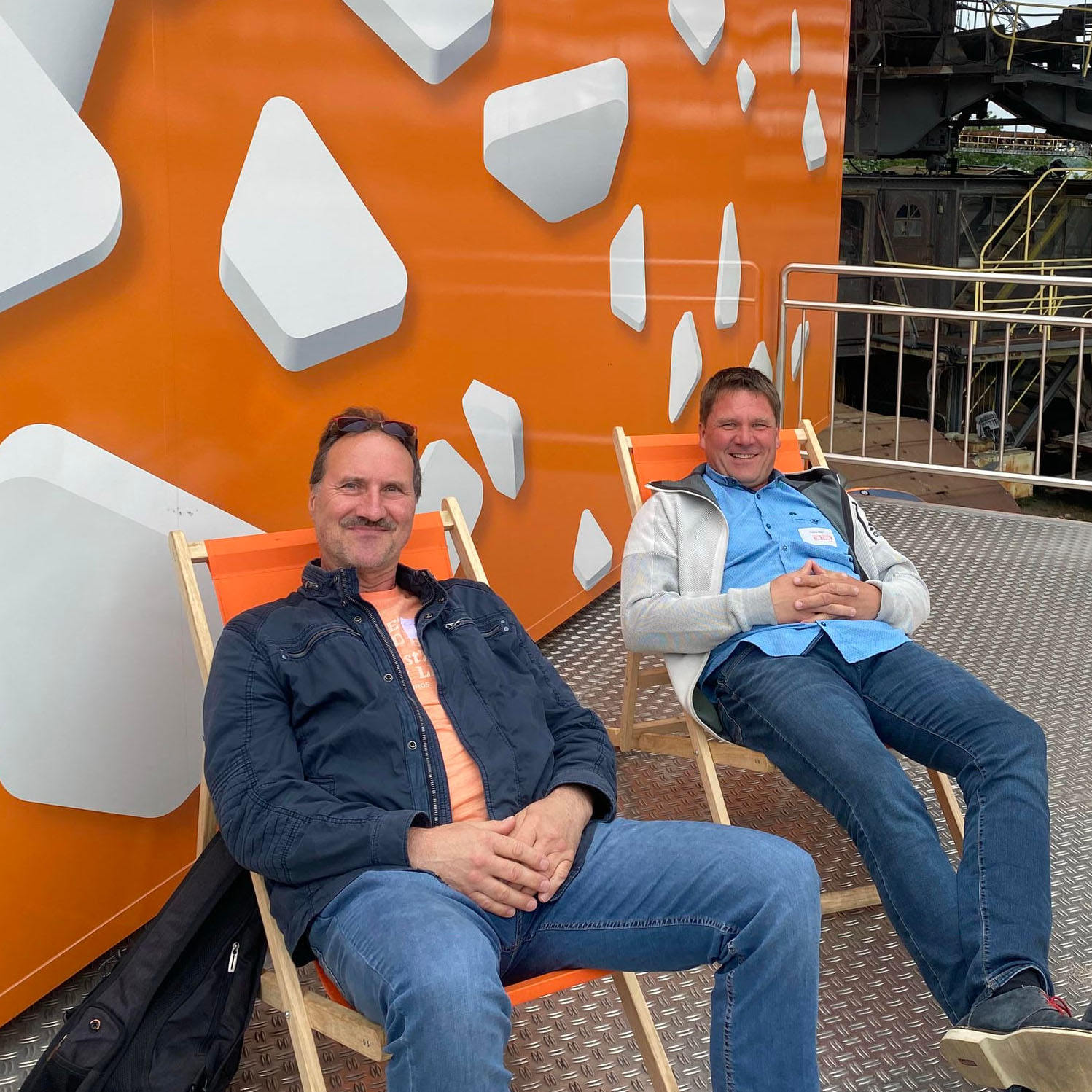 Ronny Beier und Carsten Eisold sitzen auf Stühlen vor einer orangenen Wand mit weißen, abstrakten 3D-Formen